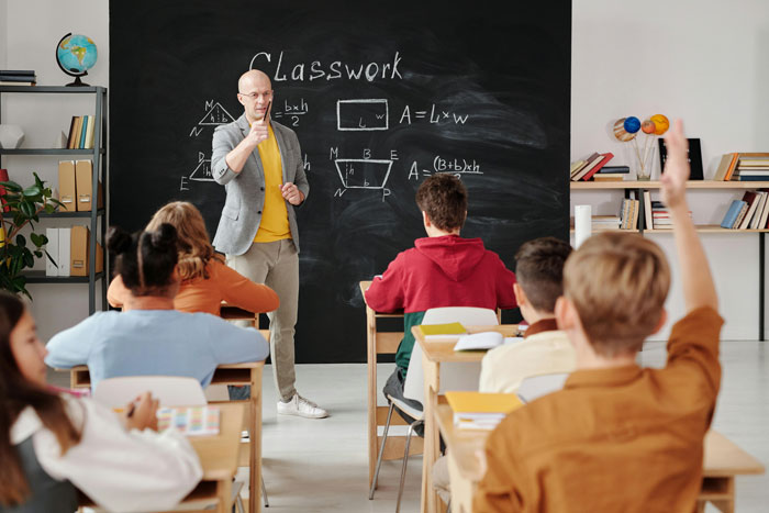 Teacher in classroom engaging students with group shaming method while students participate and raise hands during lesson.