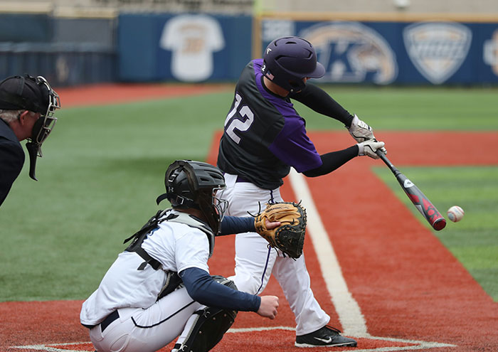 Baseball player in purple and black uniform swinging bat while catcher in white gear prepares to catch pitch on field.