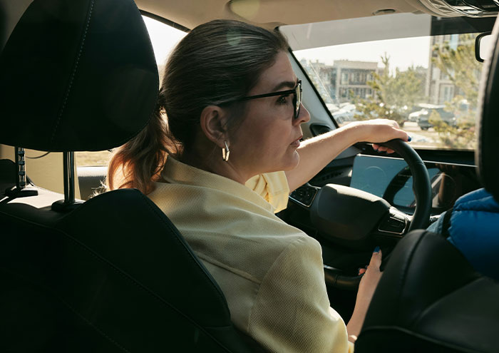 Woman driving a car, wearing glasses and gold hoop earrings, focused on the road during daytime city traffic.