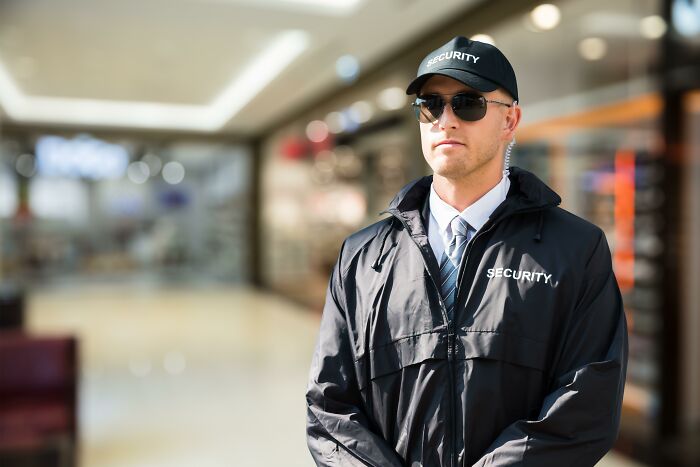 Security guard in sunglasses and black jacket standing alert in a public space, showing people in boring industries behind scenes.