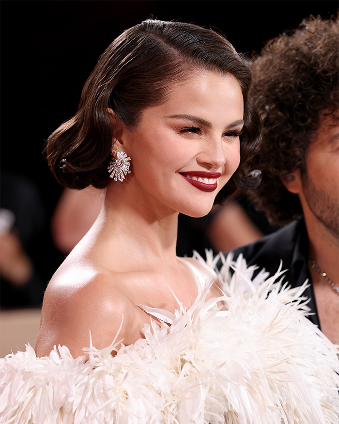 Selena Gomez attending an event in a white feathered outfit with bold makeup amid fans blaming plastic surgery transformation.