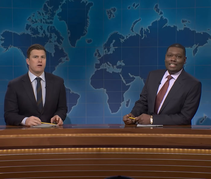 Two Weekend Update anchors on SNL set with world map backdrop, discussing a joke about ICE sparking debate.