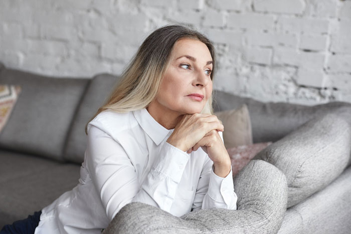 Thoughtful middle-aged woman in white shirt sitting on couch reflecting on son marrying girlfriend after high school advice.
