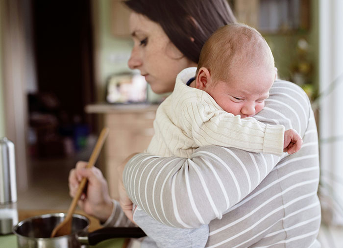 Woman holding baby while cooking in kitchen, illustrating the challenges of a man insisting his wife doesn&rsquo;t do anything at home.