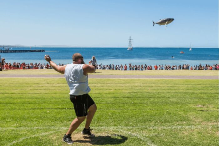 Man participating in a strange tradition of throwing a fish near the sea during a public event with spectators.
