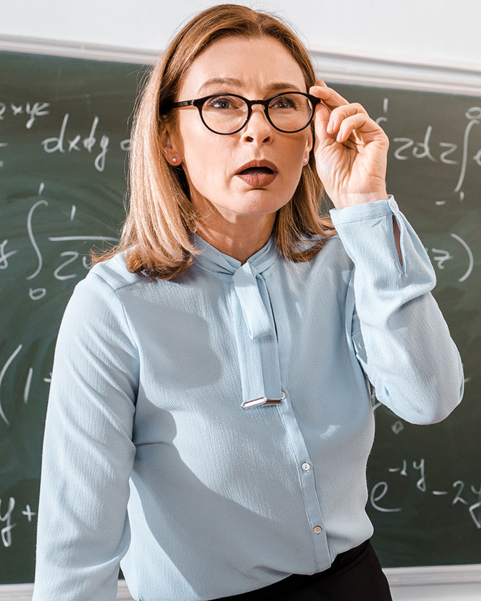 Female teacher in glasses looking surprised in a classroom, reacting to a mom struggling to contain her rage. Female teacher in glasses looking surprised in a classroom, reacting to a mom struggling to contain her rage.