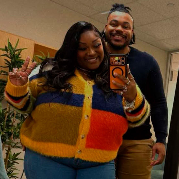 TikTok influencer posing with partner, capturing a mirror selfie indoors in colorful sweater and dark shirt.