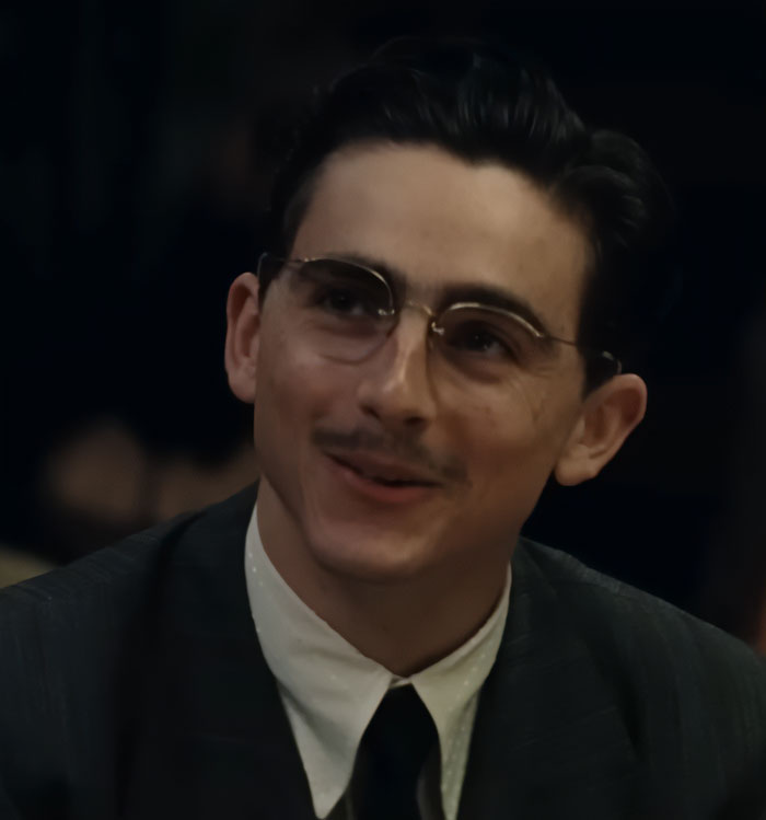 Young man wearing glasses and a vintage suit, smiling softly in a dimly lit setting related to Timothée Chalamet tone deaf message. Young man wearing glasses and a vintage suit, smiling softly in a dimly lit setting related to Timothée Chalamet tone deaf message.
