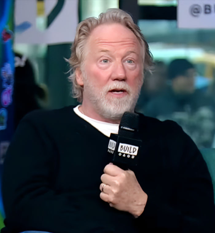 Man with gray hair and beard holding microphone during an interview about Timothy Busfield case audio revealing accusers denying claims.