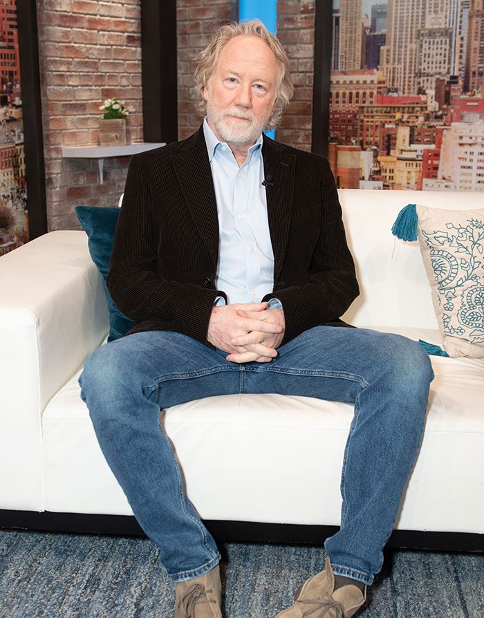 Timothy Busfield sitting on white couch in casual outfit amid discussion of case twist and audio revealing accusers denying claims.