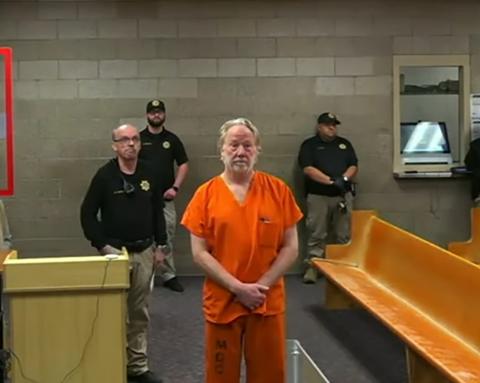 Man in orange prison uniform standing in a courtroom with officers nearby during Melissa Gilbert&rsquo;s letter case.