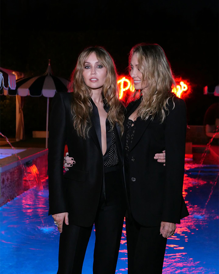 Miley Cyrus and a companion in black outfits at a nighttime red carpet event with illuminated pool background. Miley Cyrus and a companion in black outfits at a nighttime red carpet event with illuminated pool background.