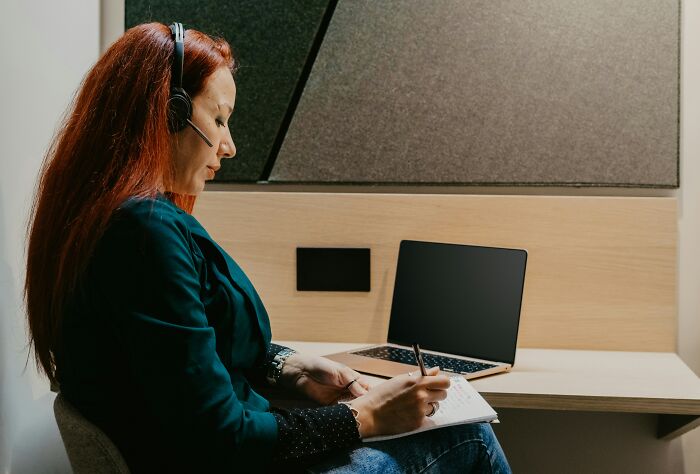 Woman with headset writing notes at a desk with laptop, illustrating stories about worst coworkers making work a nightmare