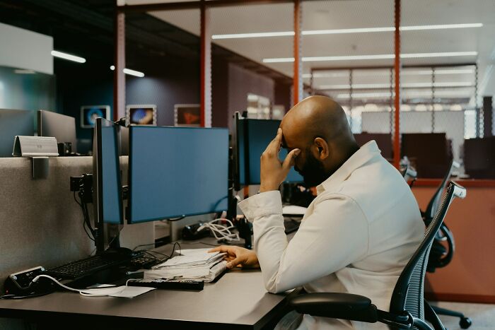Man stressed at office desk with multiple computer monitors, illustrating overrated adult goals people chase wasting time.