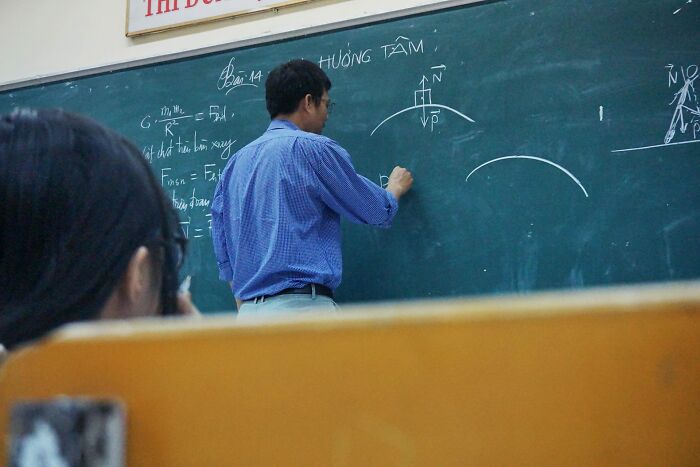 Teacher writing physics formulas on chalkboard in high school classroom during a controversial lesson discussion.