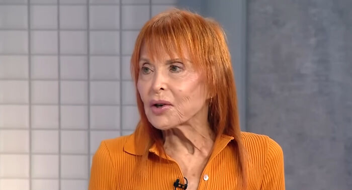 An older woman with red hair wearing an orange top, speaking during an interview about meeting idols in real life.