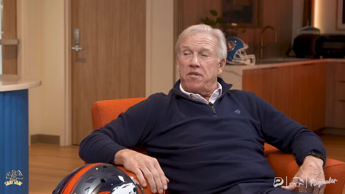 Older man in a navy sweater sitting in an orange chair with a Denver Broncos helmet nearby, sharing regret after meeting idols.