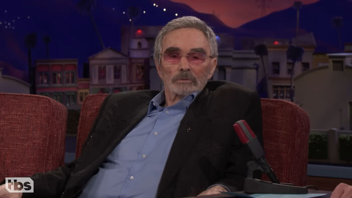 Actor wearing tinted glasses and a black blazer sitting on a talk show set about meeting idols in real life regrets