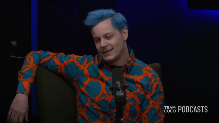Man with blue hair and vibrant orange and teal shirt speaking into a microphone during a Team Coco podcast session.