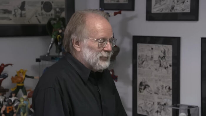 Older man with glasses and beard smiling inside a room filled with comic art and collectible figures, meeting idols moment.