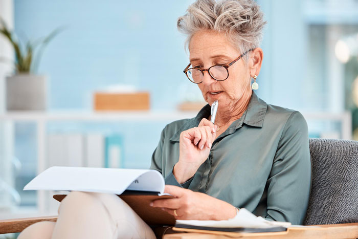 Older woman wearing glasses, seated and thoughtfully reviewing documents with pen near mouth in bright room.
