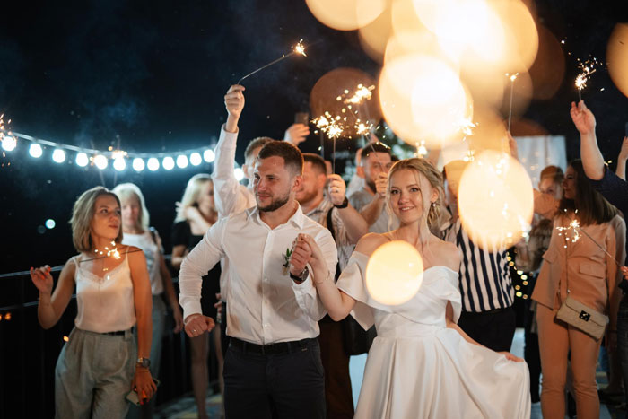 Bride in white dress holding hands with groom during nighttime wedding celebration with guests and sparklers.