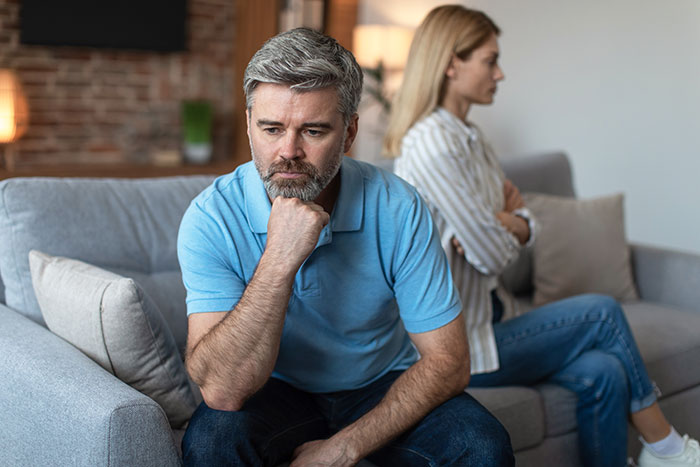 Middle-aged couple sitting apart on couch, husband looking thoughtful while wife appears upset after open marriage discussion.