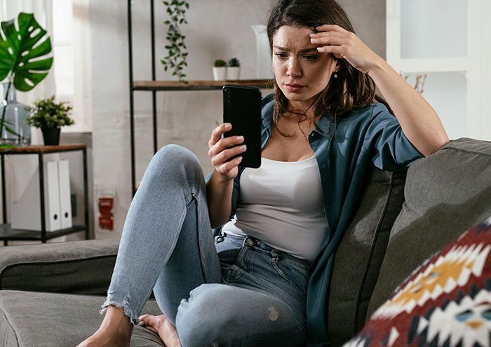 Woman sitting on couch looking stressed while holding a smartphone, with plants and shelves in the background.