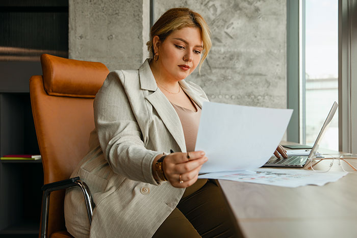Young mom reviewing documents at desk, appearing thoughtful and focused while working on her laptop near a window. Young mom reviewing documents at desk, appearing thoughtful and focused while working on her laptop near a window.