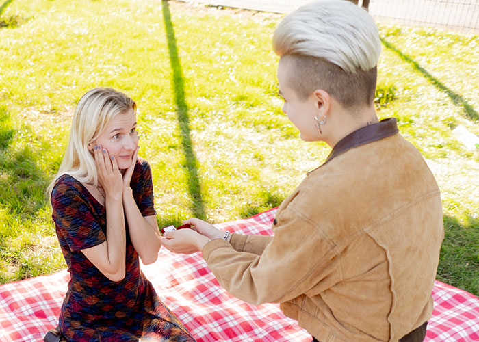 A person proposing outdoors on a red plaid blanket with a surprised fiancée showing an emotional reaction. A person proposing outdoors on a red plaid blanket with a surprised fiancée showing an emotional reaction.