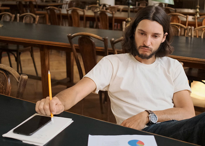 Man in white t-shirt sitting thoughtfully at a table, focused on a phone and notes while holding a pencil.