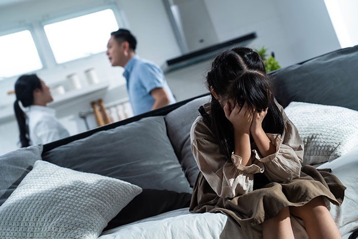 Woman covering face in distress on couch while parents argue in background, illustrating conflict over kids and trip to Italy.