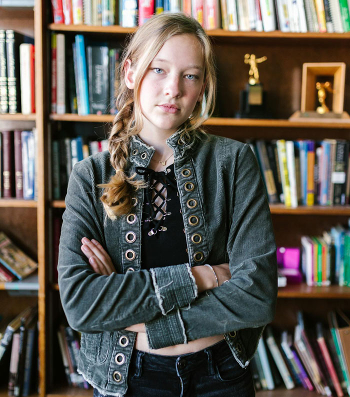 Young woman with braided hair standing with crossed arms in front of a bookshelf, reflecting on fianc&eacute; and stepdaughter conflict.