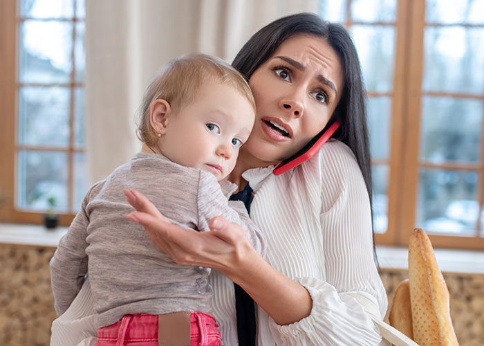 Woman holding baby and talking on phone with a worried expression about engagement party and friendship issues.