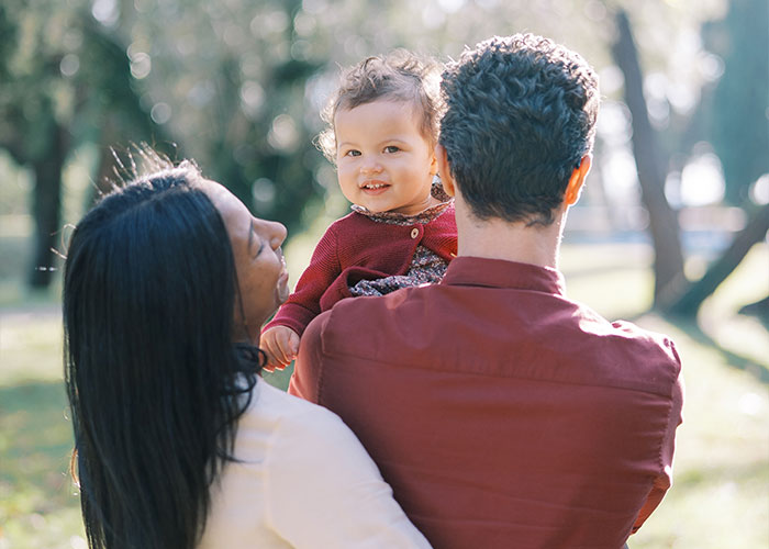 Young woman and her husband holding their smiling baby in a sunlit park, highlighting friendship and family dynamics.