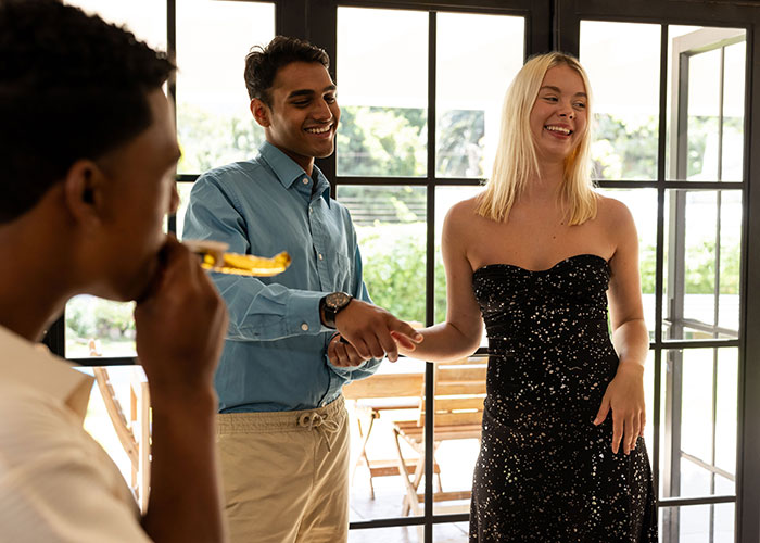 Young woman and man smiling and shaking hands at an engagement party, while another guest blows a party horn.