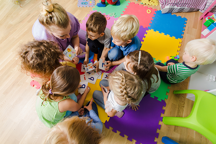 Daycare worker engaging with children on colorful mats, using educational alphabet cards in a playful learning setting.