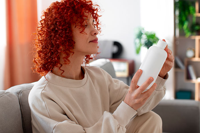 Daycare worker with curly red hair examining a white bottle, reflecting on possible passive-aggressive coworker gift at home. Daycare worker with curly red hair examining a white bottle, reflecting on possible passive-aggressive coworker gift at home.