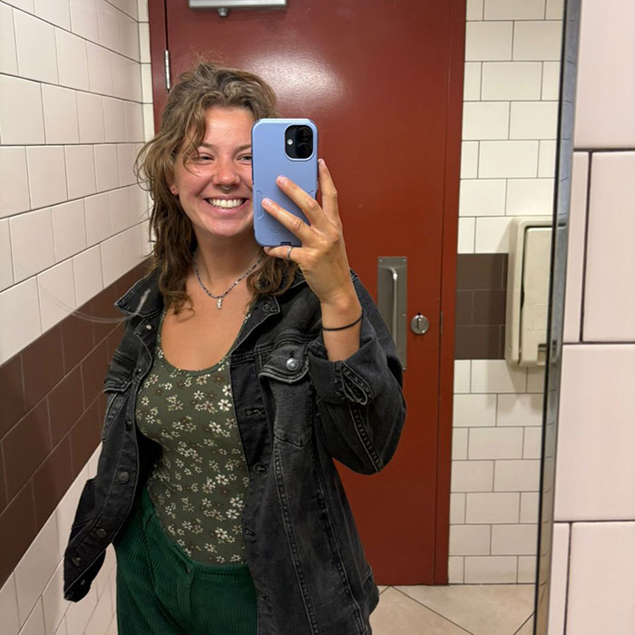 Woman taking a mirror selfie smiling in bathroom, related to breast reduction and online trolling controversy. Woman taking a mirror selfie smiling in bathroom, related to breast reduction and online trolling controversy.