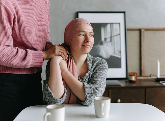 Woman with cancer wearing a headscarf sits at table holding hands with a standing person, conveying emotional support and motherhood.