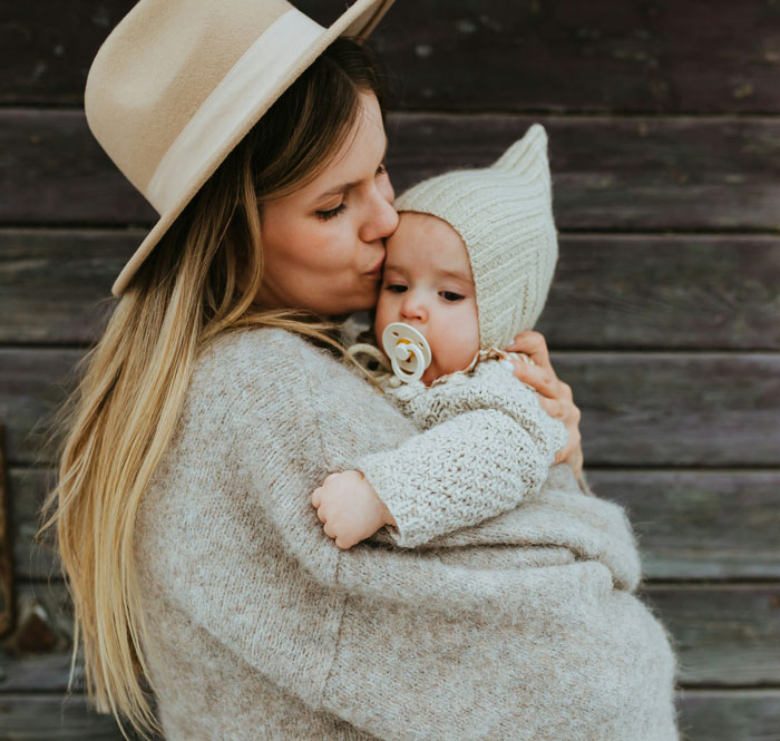 Woman wearing hat holding and kissing baby dressed in knitwear, illustrating cousin with cancer wanting to borrow baby for motherhood experience.