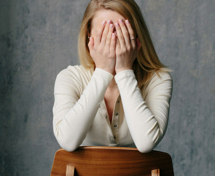 Woman in a white shirt sitting on a chair covering her face, reflecting feelings about cousin with cancer and motherhood experience.
