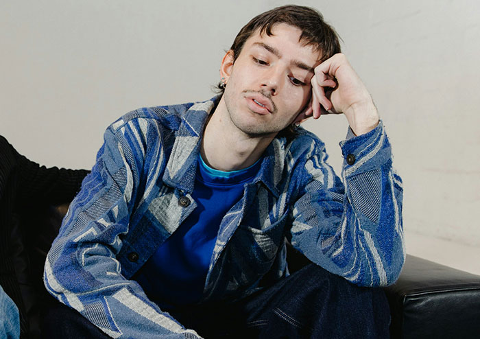 Young man in a patterned blue jacket sitting thoughtfully on a black couch, reflecting on what women obsess over.