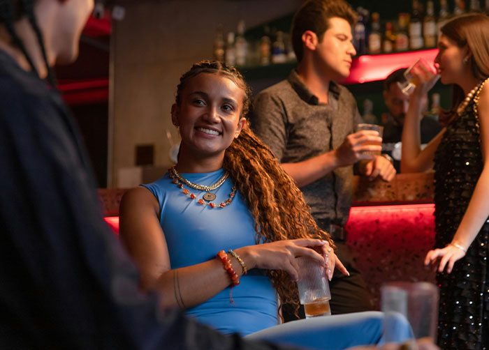 Young woman smiling and holding a drink at a bar, socializing with friends about what women obsess over for no reason.
