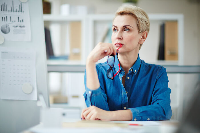 Woman in blue shirt holding glasses, thoughtfully looking away, illustrating misconceptions about women in modern times.