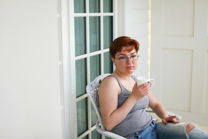 Woman with short hair and glasses sitting on a chair by a window holding a cup, reflecting on misconceptions about women.
