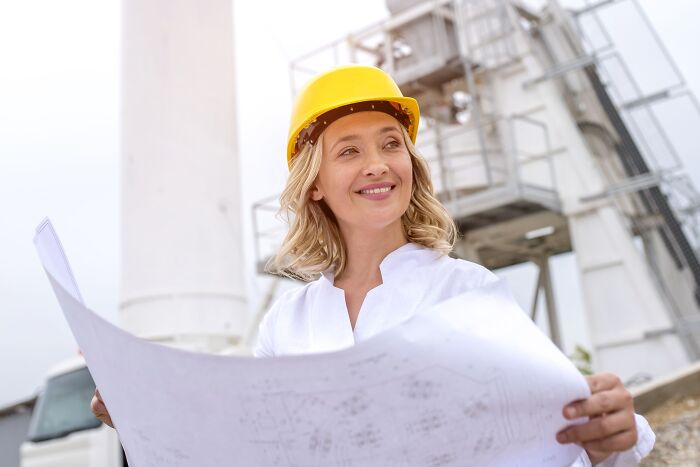 Woman engineer wearing a yellow hard hat, holding blueprints at a construction site challenging misconceptions about women.
