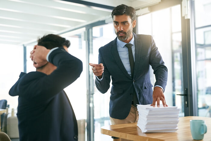 Angry businessman pointing at overwhelmed employee with large stack of documents, depicting moments people realized to quit job.