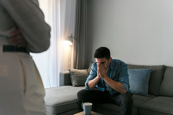 Man sitting stressed on a couch during a tense conversation with his girlfriend about a business trip.
