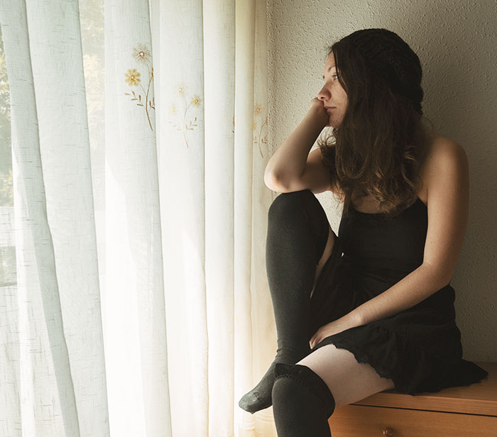 Woman in black dress and knee socks sitting by window, looking thoughtful, relating to business trip misunderstanding.
